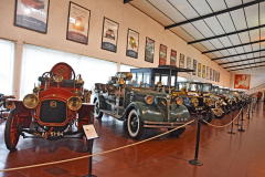 Defilé nejstarších automobilů. Na stěnách jsou plakáty příležitostných automobilových výstav muzea. Za ­požárními automobily se tyčí mohutný De Dion-Bouton 24CV