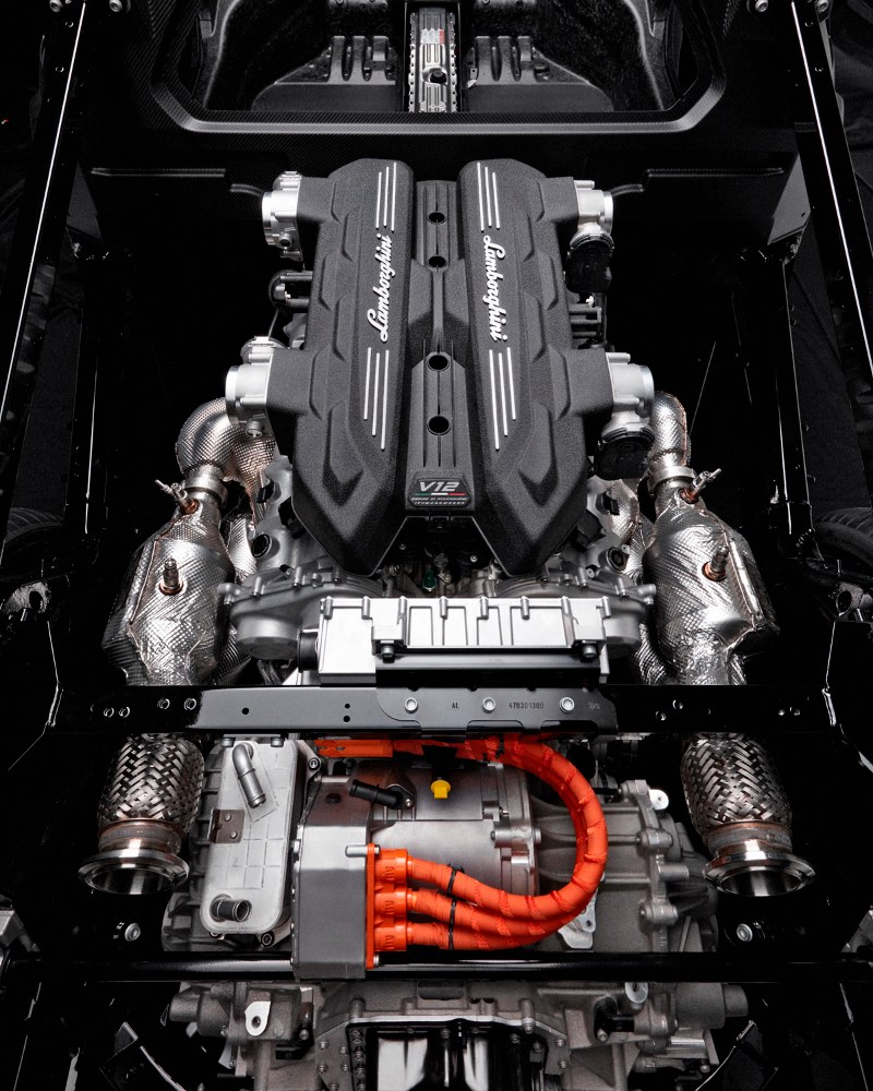 Komory sání motoru V12 vyčnívají zpod kapoty. Ve spodní části je vidět napříč uložená převodovka, jejíž součástí je elektromotor